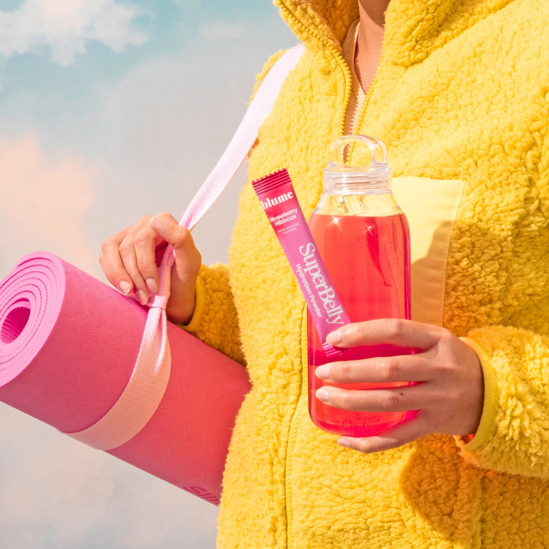 A person in a yellow fleece jacket holds a rolled pink yoga mat, a packet labeled Blume SuperLatte, and a pink drink in a clear bottle. The background is a soft, cloudy sky.